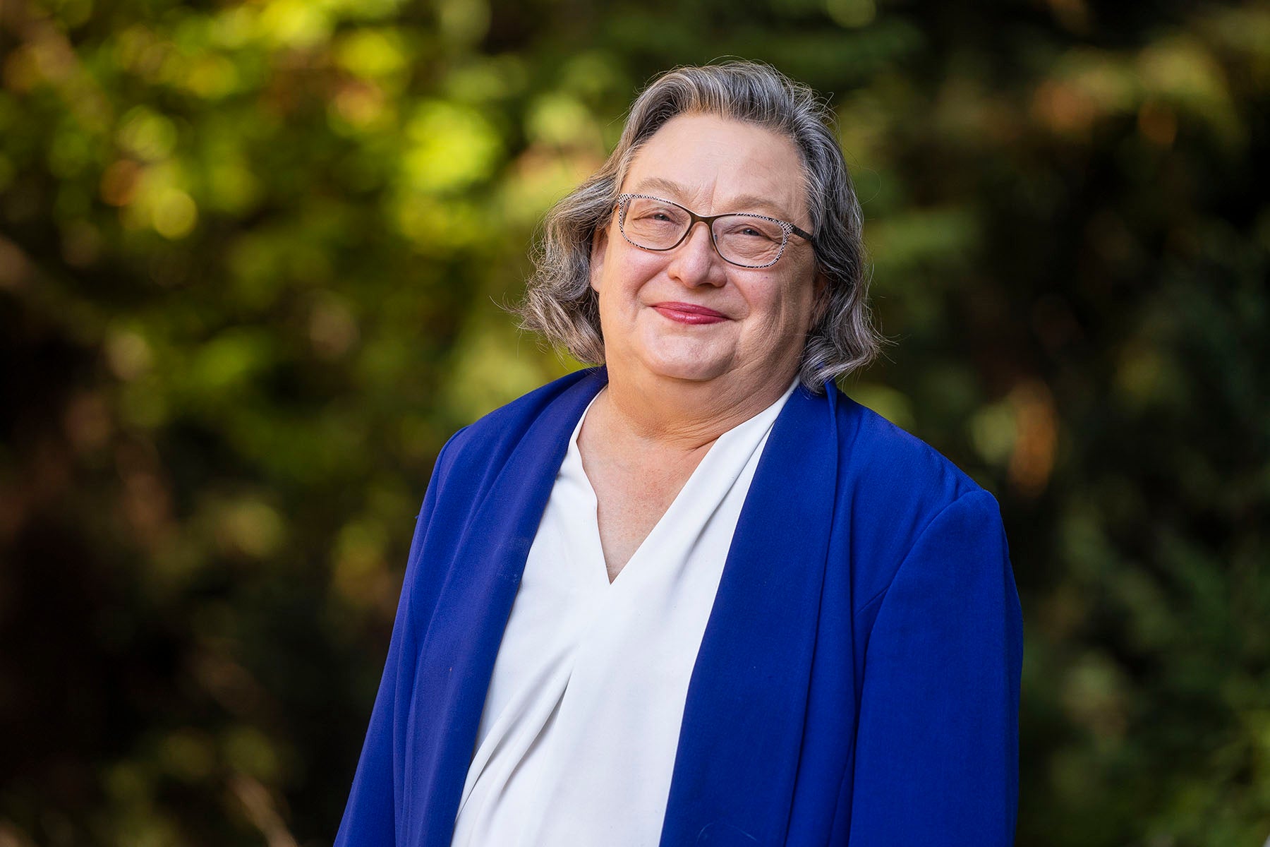 Portrait of Chancellor Larive, who smiles at the camera wearing a blue blazer and white blouse, against a backdrop of greenery.