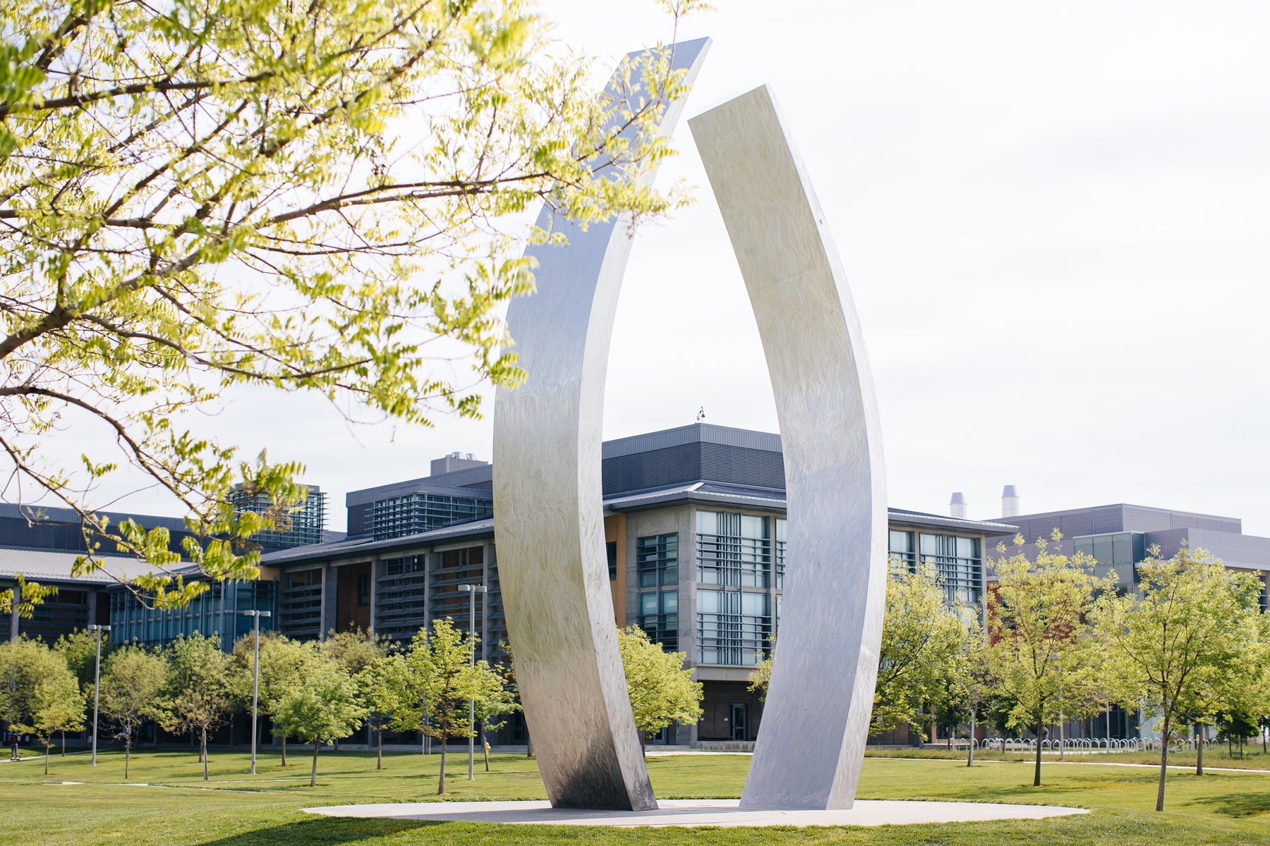 A giant metal sculpture with two curving vertical pieces, in a parklike setting with buildings in the background