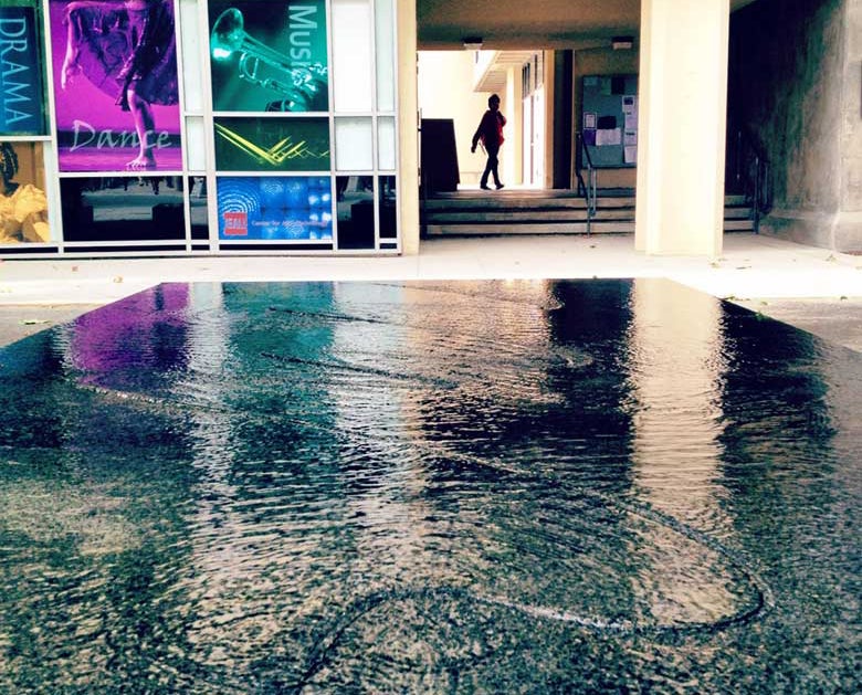 A flat fountain covered with water with curving linear patterns, with a breezeway and a person walking in the background