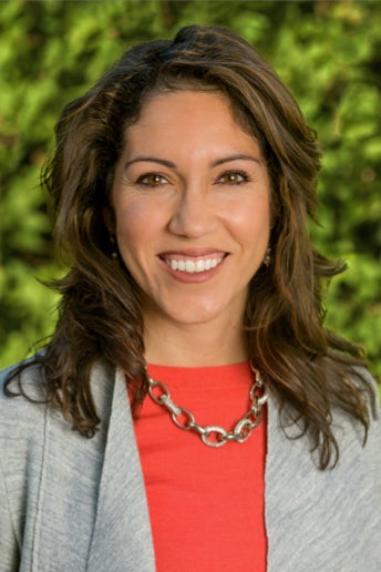 Maria Anguiano, woman with wavy shoulder-length hair in a gray jacket and red top with a necklace, smiling