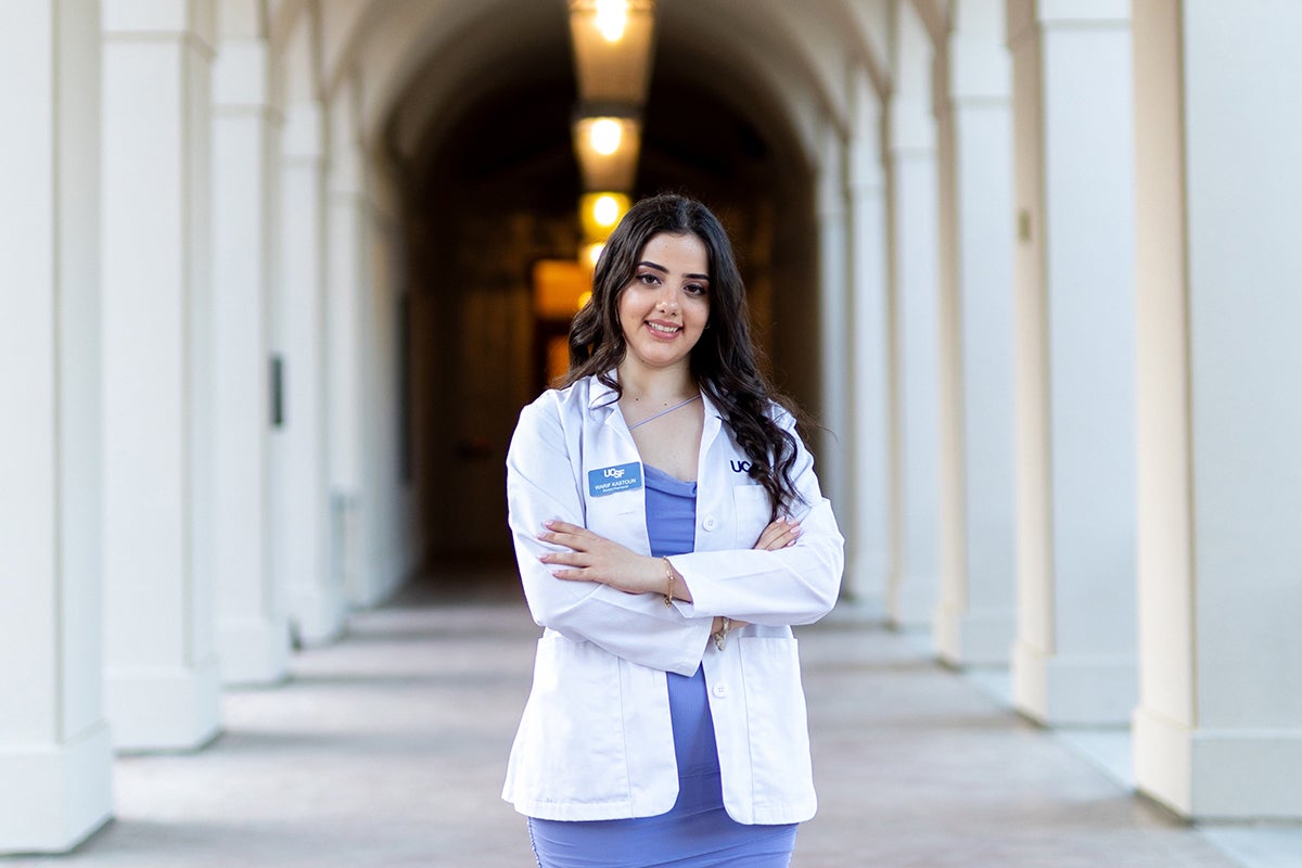 A young woman with long dark hair in a white coat poses surrounded by columns, arms crossed