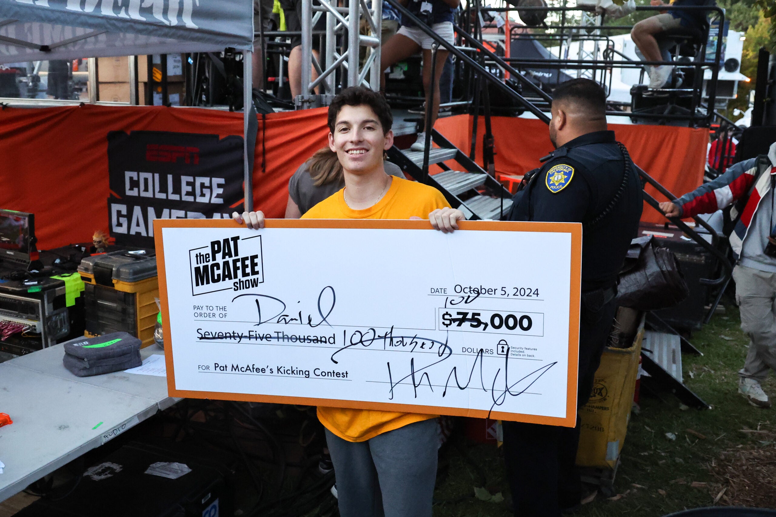 A young man in a Cal gold T-shirt holds up a check with 75,000 crossed out for 100,000 behind the GameDay set