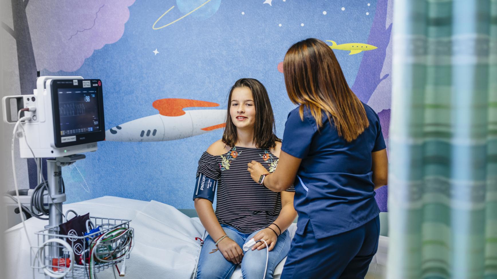 A nurse listens to the heartbeat of a young teen sitting up on a bed in a room with colorful spaceship wallpaper