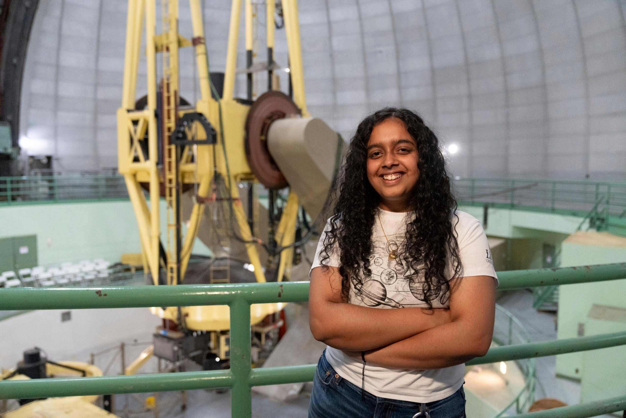 A student with long hair smiles in front of a telescope inside an observatory dome