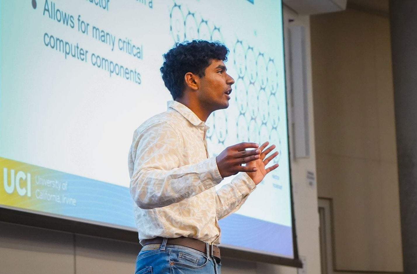 A young man in a white-collared shirt gives a talk in front of a large screen