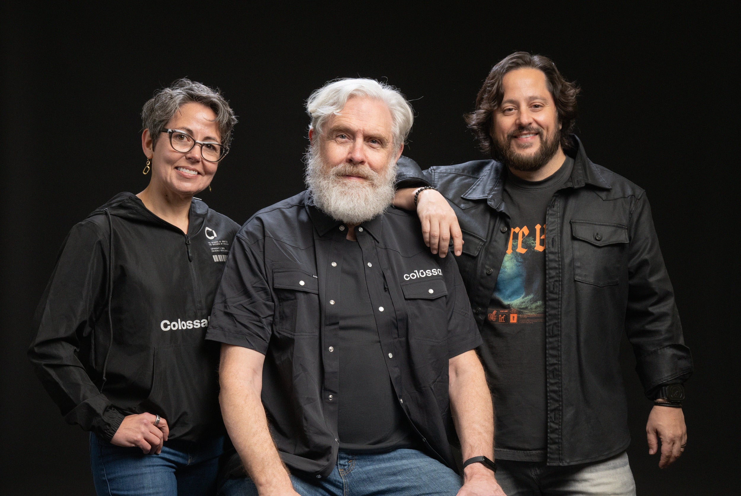 Three people stand together wearing black tops and jackets and vests on a black background