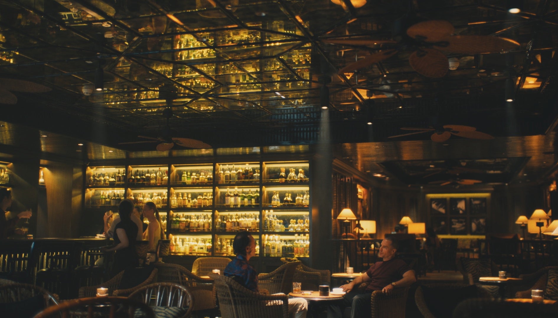 Two characters sit in a bar lounge with many mirrors and yellow light in a scene from The White Lotus