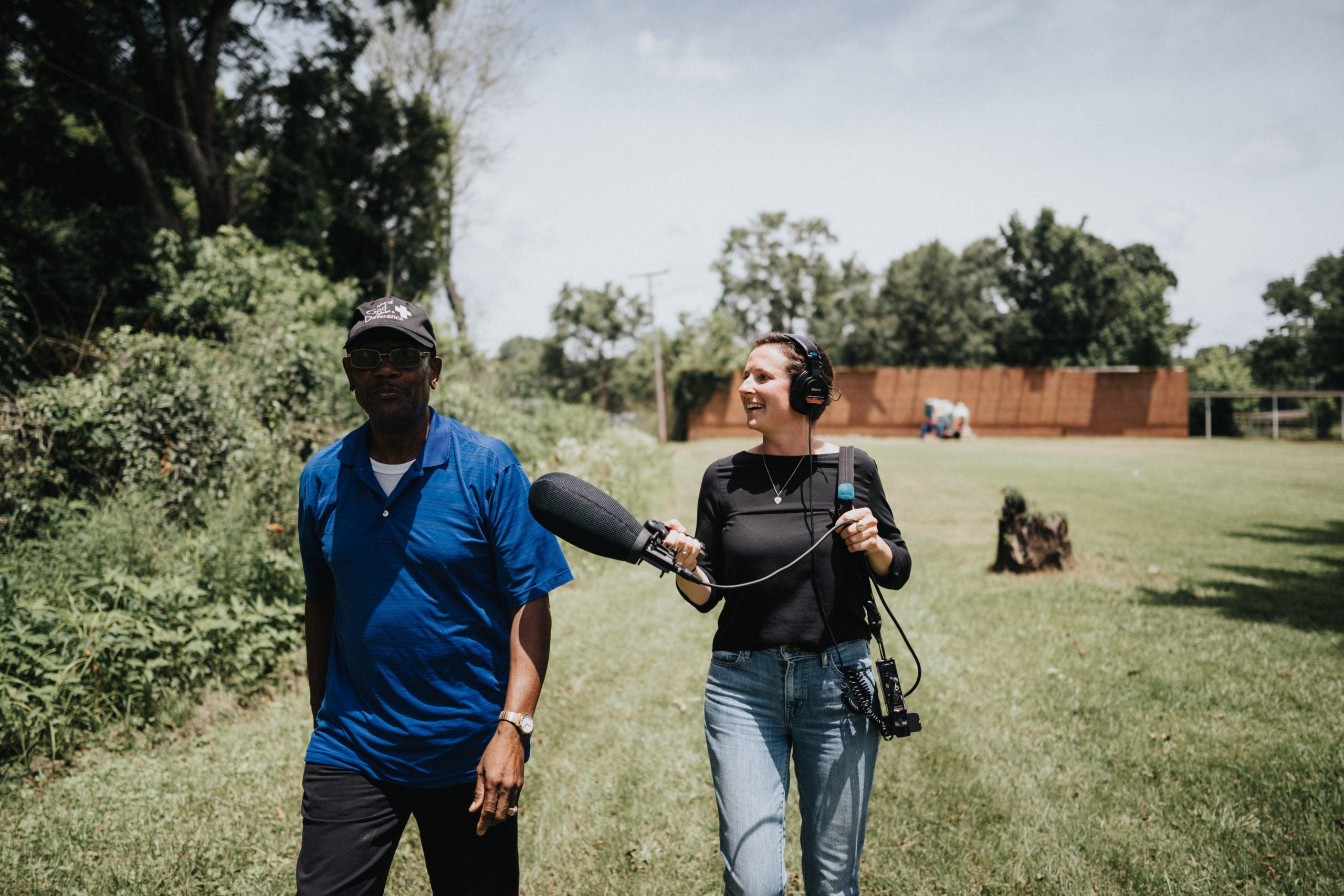 A woman trails slightly behind a man with a giant microphone recording him as he walks through a yard