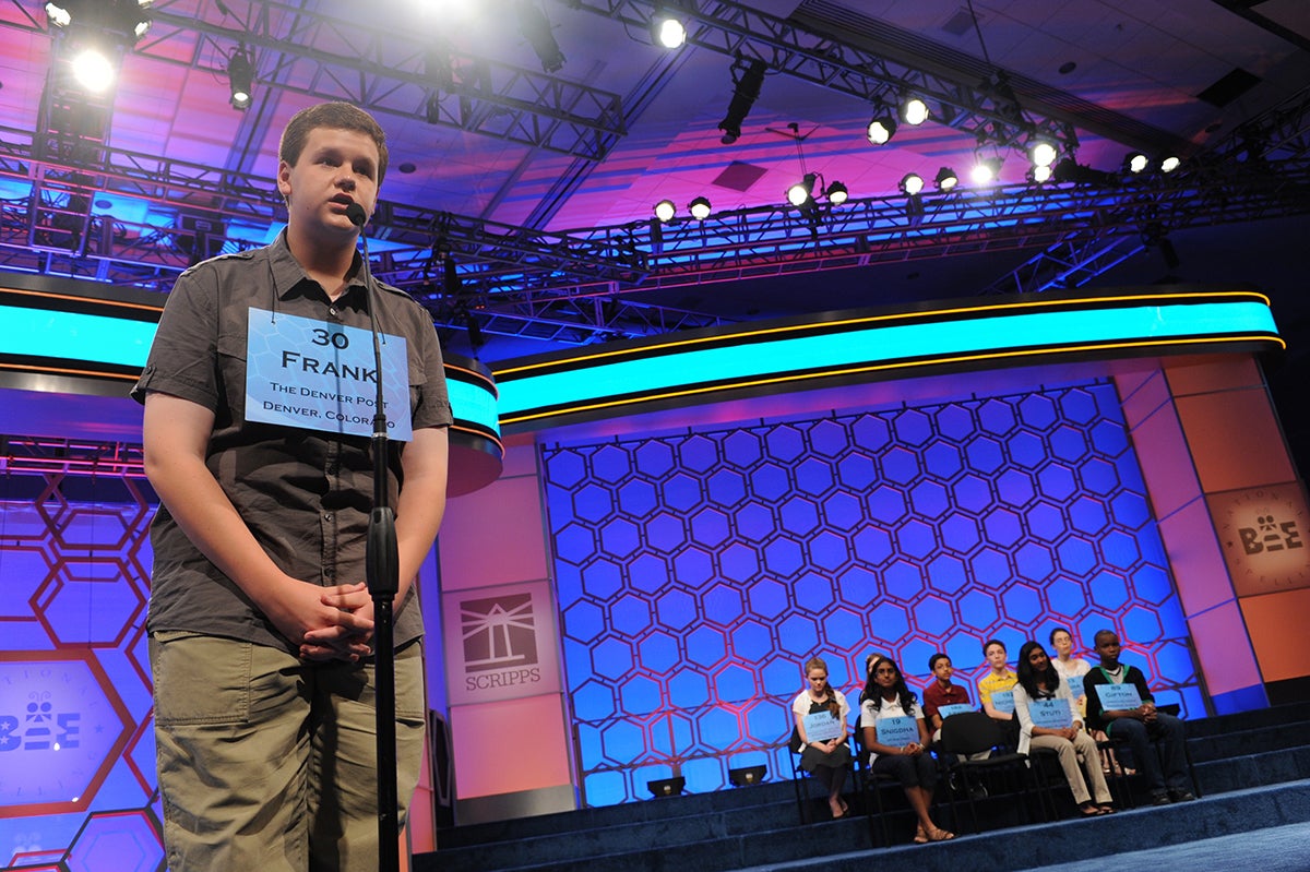 A teenager at a microphone during a spelling bee, other contestants visible in the back