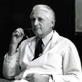 A man with white hair in a doctor's coat and tie holds his glasses