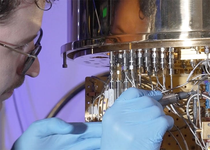 A student wearing glasses and blue gloves works closely on a complicated, gold-colored machine