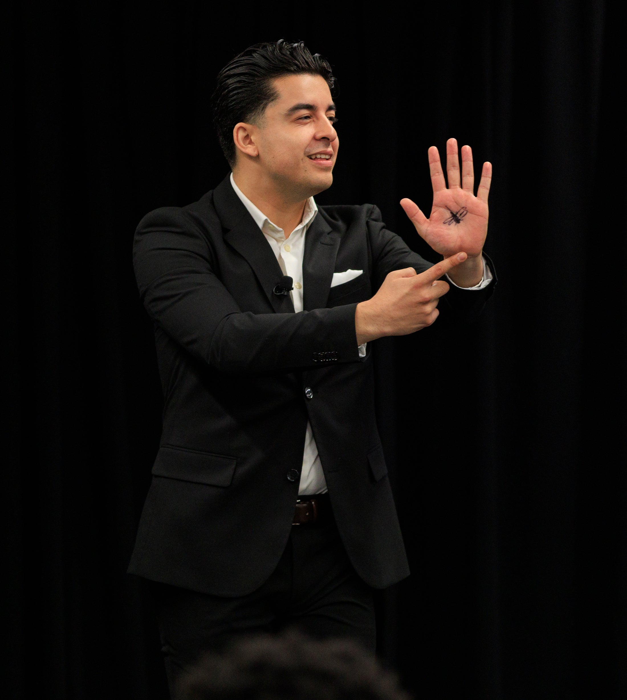 A young man in a black suit jacket holds up his hand, with a drawing of a mosquito on his palm
