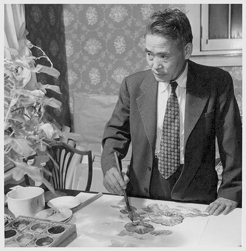 A man in a suit and tie standing at a table painting with brush and ink