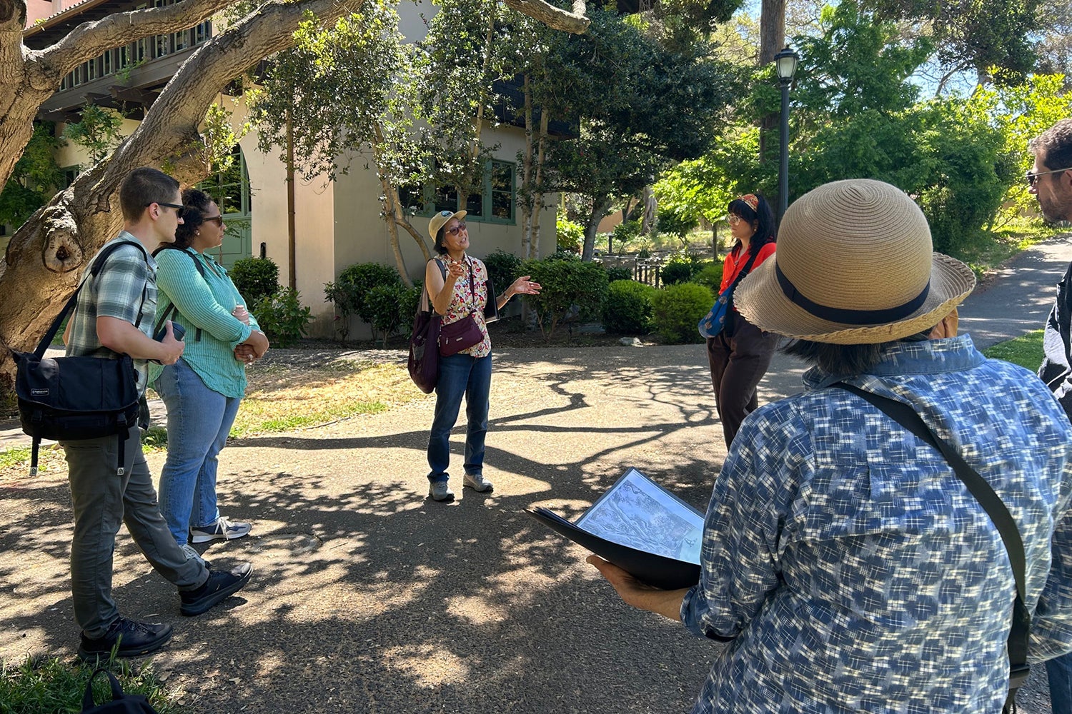 A woman in a straw hat talks to a group of people under an oak tree on a campus tour