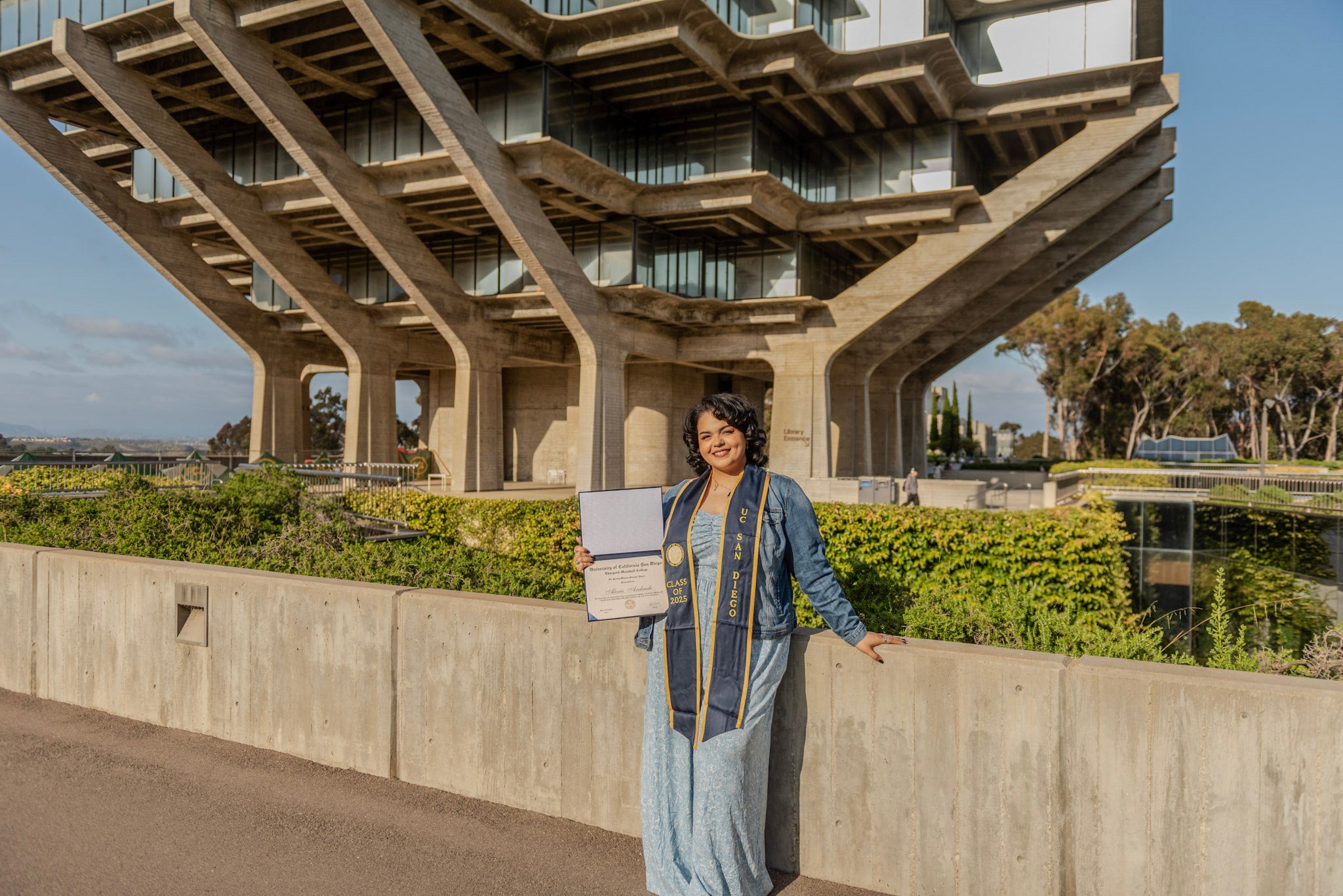 A woman in light blue dress and dark blue stole shows her diploma while standing on an outdoor walk at UC San Diego, with Geisel Library and the sea behind her