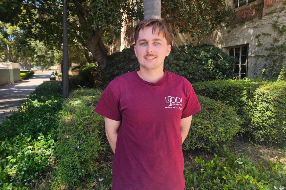 Young man with mustache smiles at camera in front of leafy area on UCLA campus