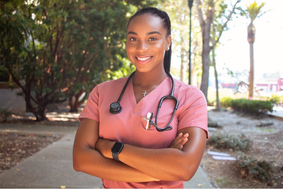 A woman in a pink nursing scrub with a stethoscope around her neck smiles at the camera, arms crossed