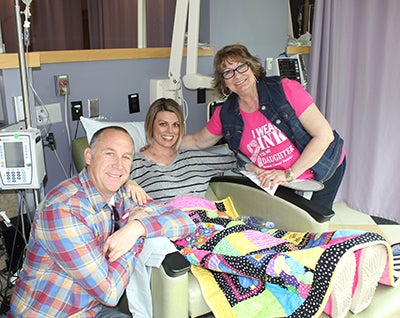 A woman in a hospital bed smiles with a woman leaning over her smiling and a man kneeling on the other side, smiling