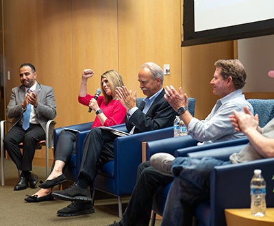 Four people in chairs in a wood-paneled room, several applaud and a woman pumps her first