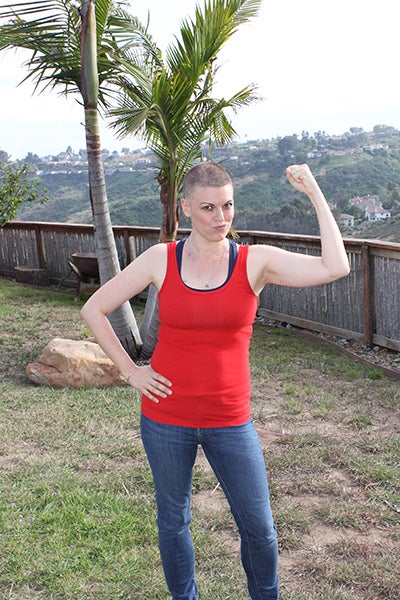 A woman with a shaved head flexes her bicep with her other hand on her hip, standing outside in workout clothes