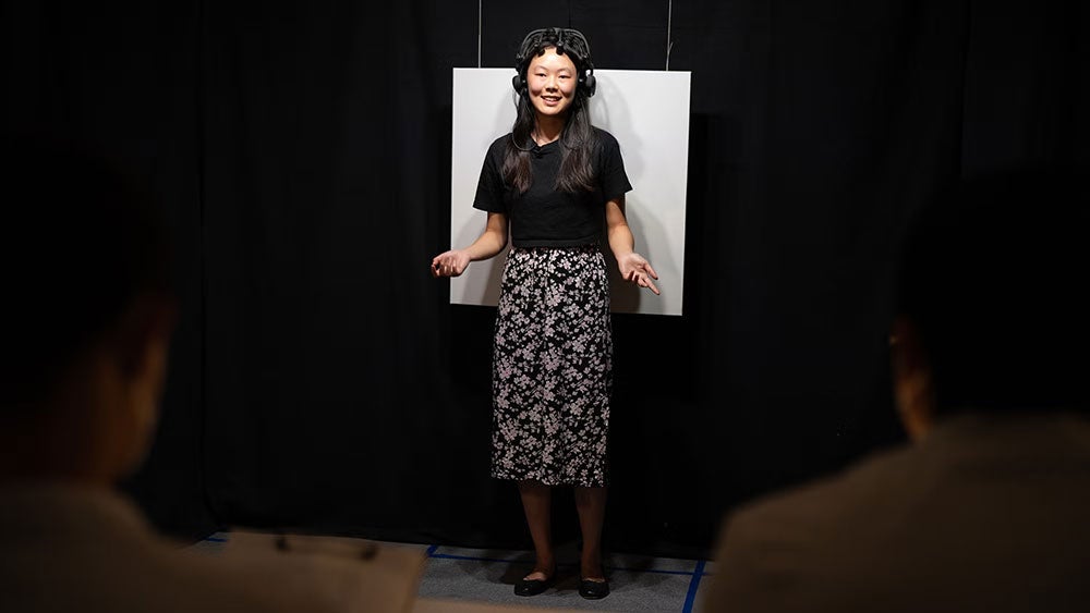 Veronica Then speaks in front of 2 people in a dark lab, standing in front of a white screen, wearing an electronic measuring device on her head.