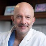 A bald man with a mustache in a white doctor's coat, smiling