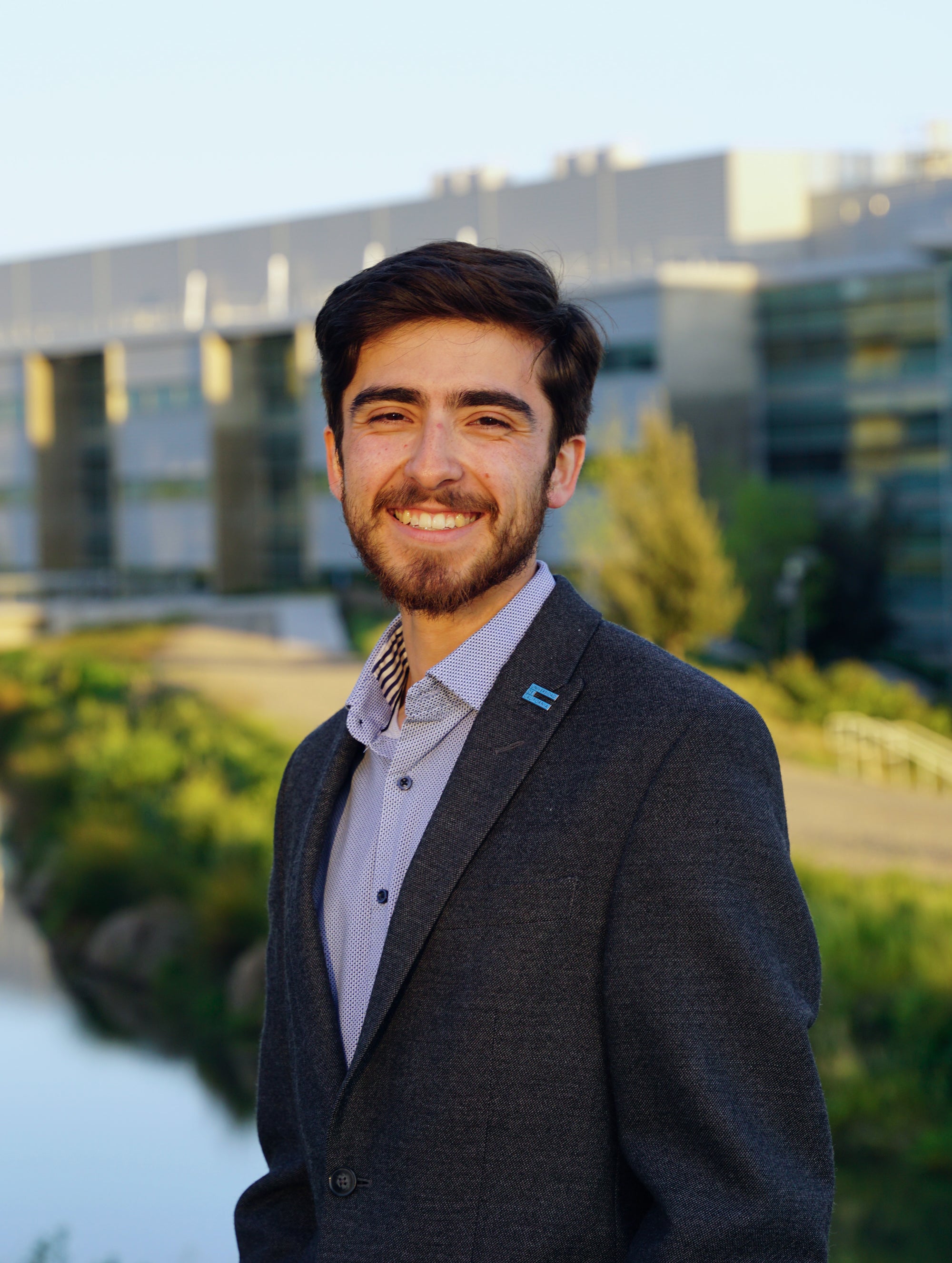 A young man with a closely-trimmed beard in a blue shirt and dark blue jacket smiles