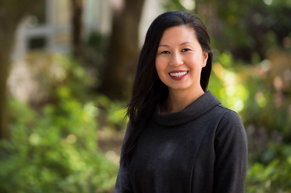 A woman in a sweater with long black hair smiles outside in front of out-of-focus shrubbery
