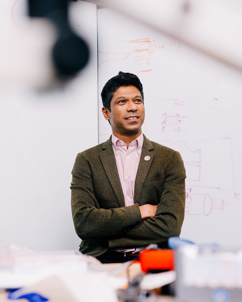 Man in a pink dress shirt and brown suit jacket with arms crossed in front of whiteboard