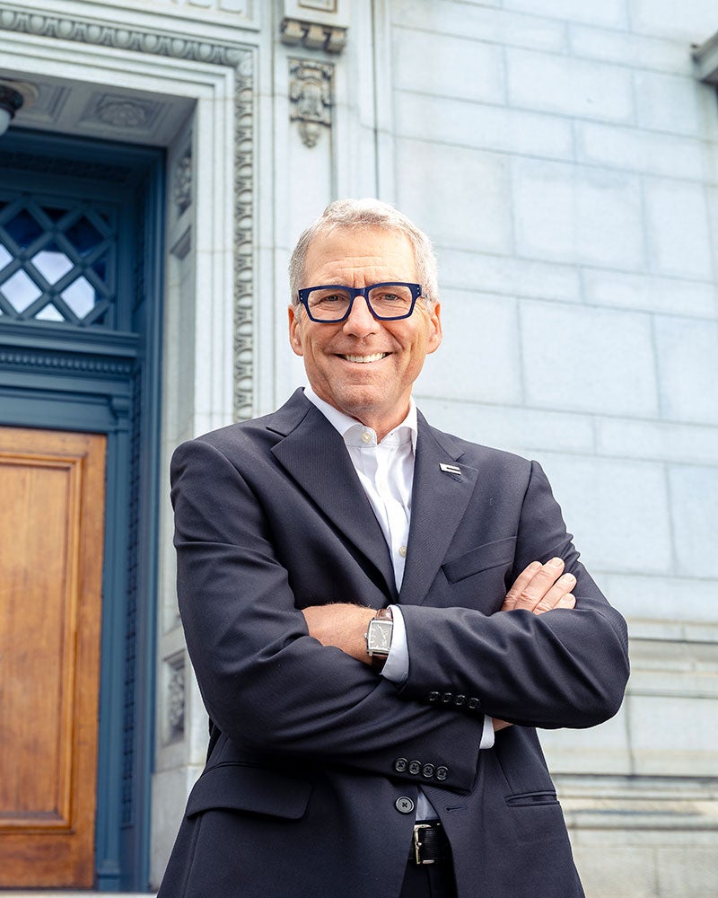 James Milliken smiles at the camera in a suit jacket with his arms folded over his chest, in front of a marble building with a blue door.