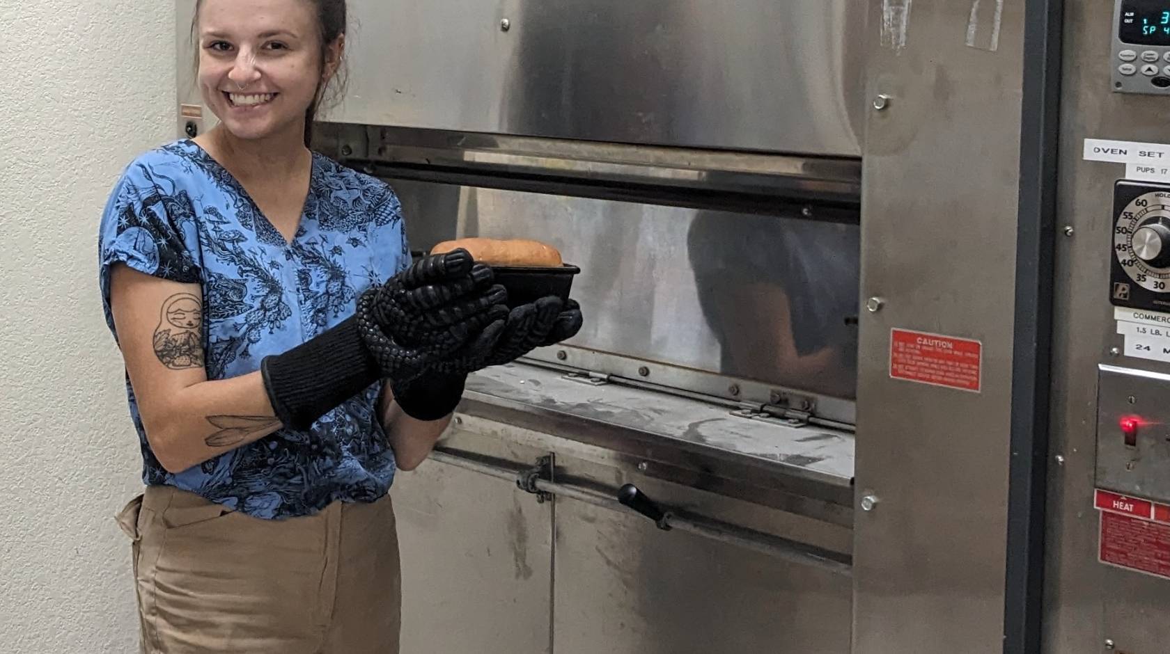 A woman holds a loaf of bread and smiles next to an oven