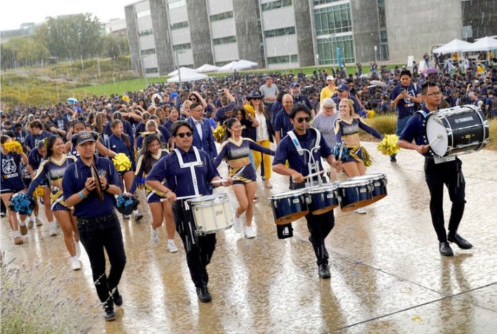 A marching band leads a large procession down a wide campus walkway in a downpour