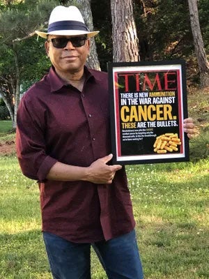 Mel Mann, wearing sunglasses and a fedora, holds a framed copy of a TIME magazine cover announcing the release of Gleevec.