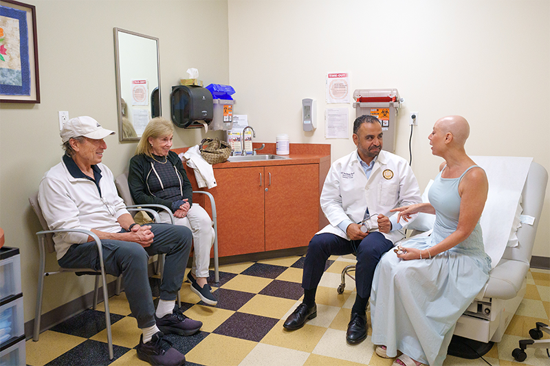 A woman without hair on her head from chemo in a pale blue dress holds her doctor's hand as he speaks to her in a room, her parents looking on