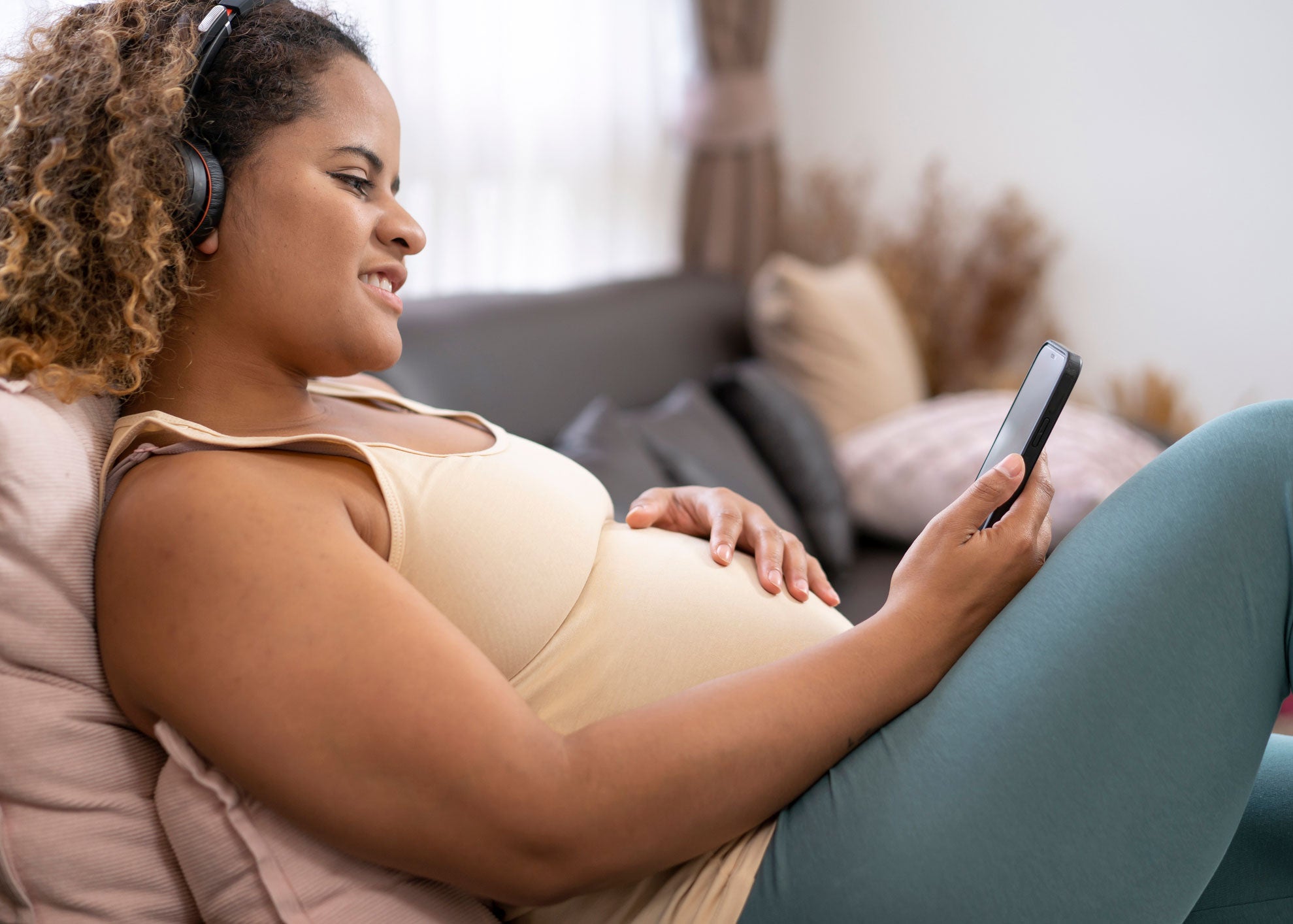 A woman reclines on a couch wearing headphones, looking at a phone, smiling, with her hand on her pregnant belly