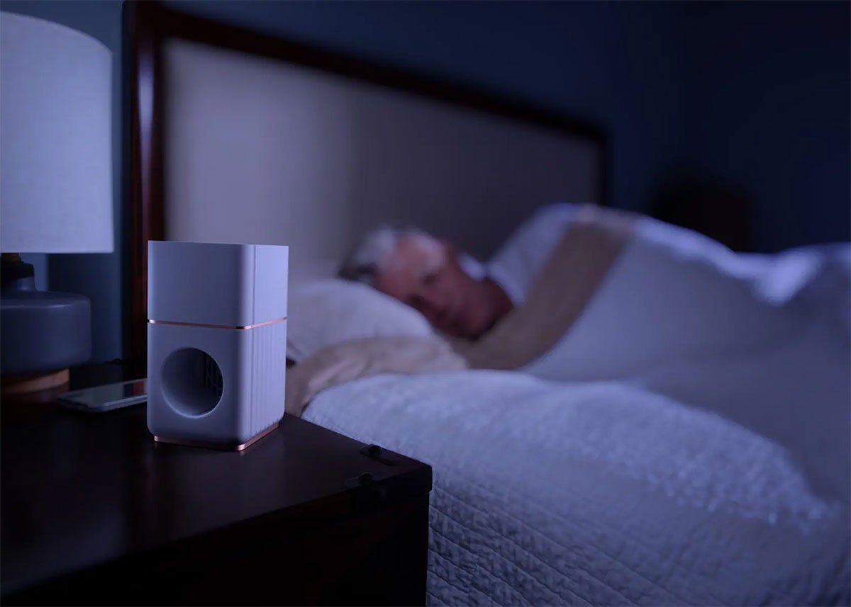 A white device about the size of a coffee mug sits on a bedside table next to an older gentleman asleep in bed