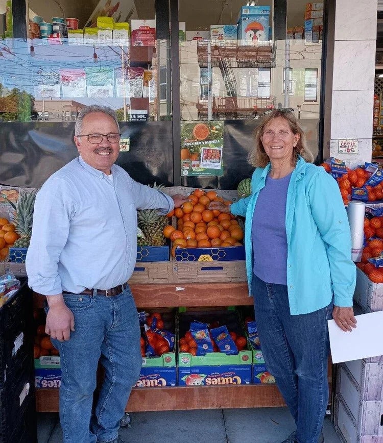 A man and a woman stand outside a market, flanking some oranges in a stand
