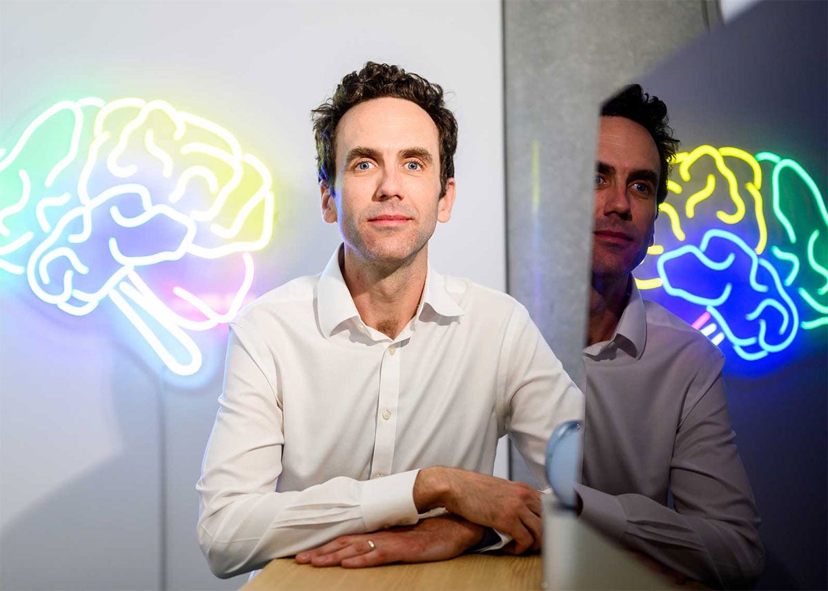 Simon Little looks at the camera, sitting in front of a large wall art that's a neon tube rendering of the human brain