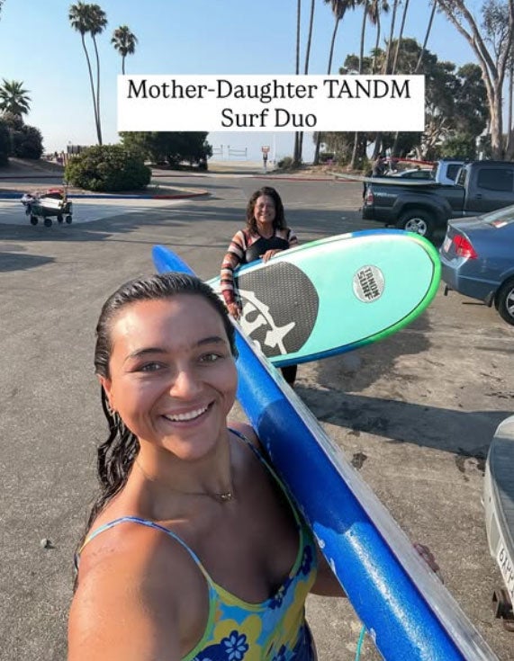 JuJu Clark and her mom pose for a selfie with surf boards in the parking lot at the beach. Text over the image reads "Mother-Daughter TANDM Surf Duo"