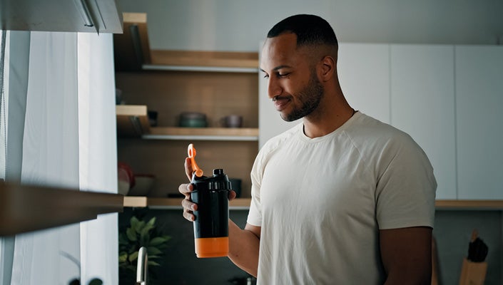 A healthy-looking man smiles at a shake he's holding in his hand, standing in his kitchen