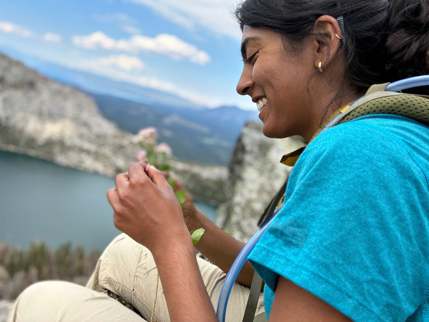 A person wearing a turquoise shirt and backpack sits on a rocky mountainside overlooking a lake and distant peaks, smiling while examining a small pink wildflower in their hands. The bright sky and expansive view suggest a high-elevation hiking or fieldwork setting.