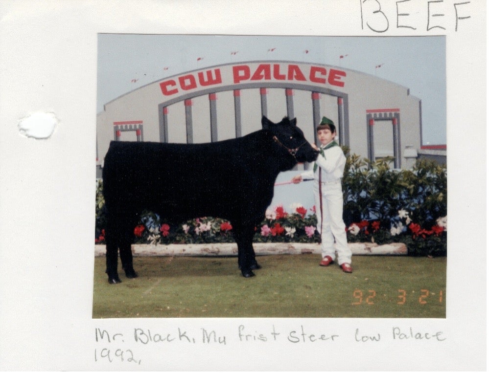 In a photo from the 90s, a young girl in a 4-H costume stands with a black steer in front of a backdrop that says Cow Palace