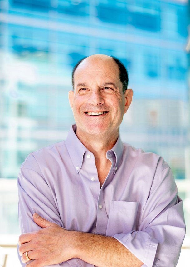 David Julius looks off to the side of the camera, smiling, wearing a purple button up shirt with his arms folded over his chest.