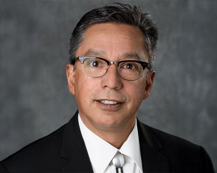 Hector Gonzalez smiles at the camera against a gray studio background wearing a black suit and a bolo tie