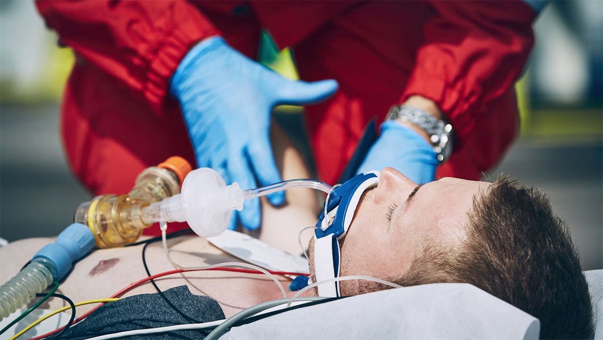 A paramedic wearing blue gloves and red protective gear provides emergency care to an unconscious man lying on a stretcher, who has a breathing tube and medical monitoring wires attached to his chest.