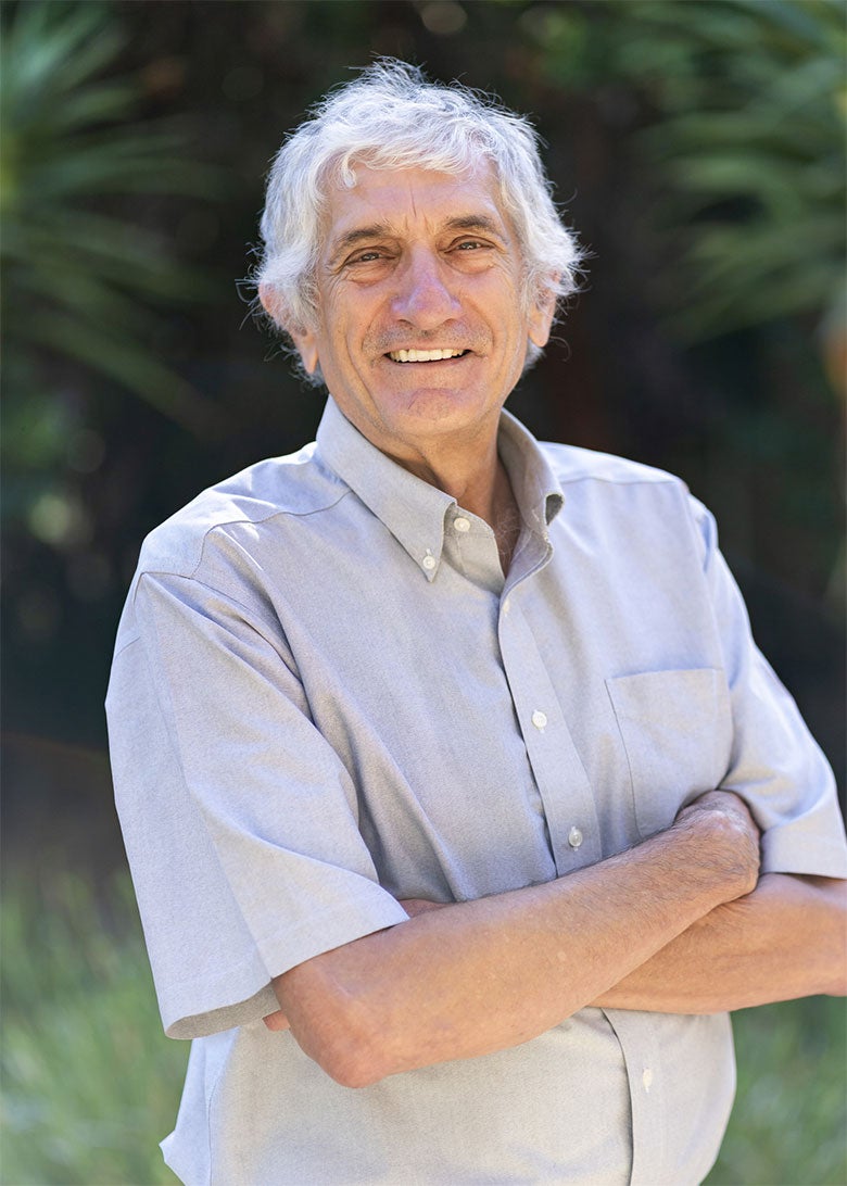 John Martinis smiles at the camera with his arms crossed over his chest, standing in front of vegetation.