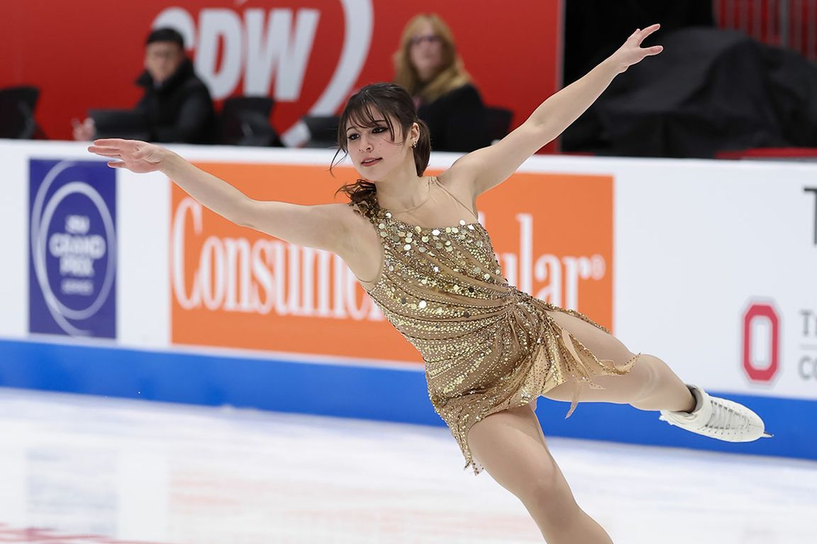 A woman in a beige skating outfit extends her arms while skating