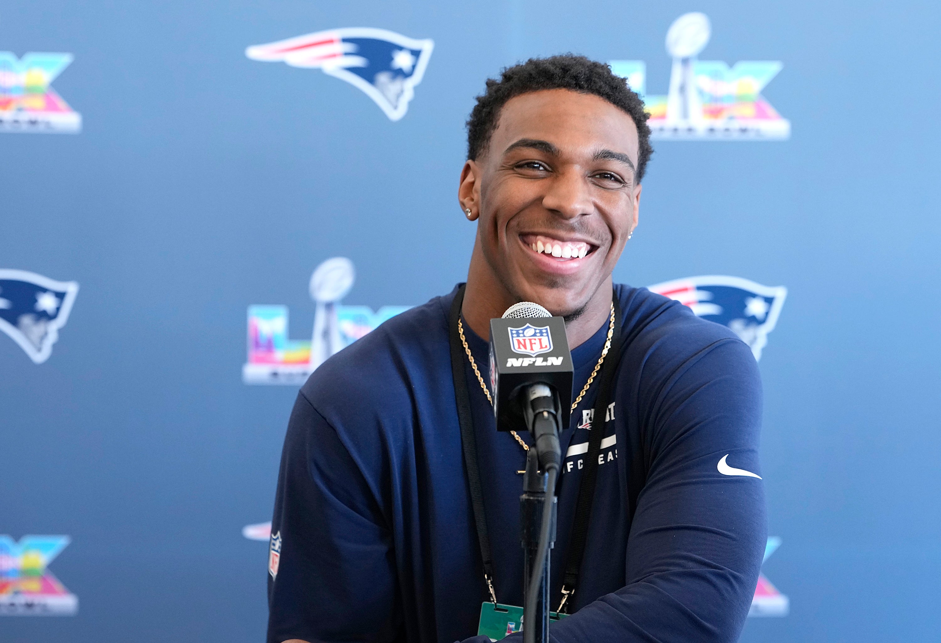 A man smiles at a podium with Super Bowl logos and the New England Patriots logo behind him