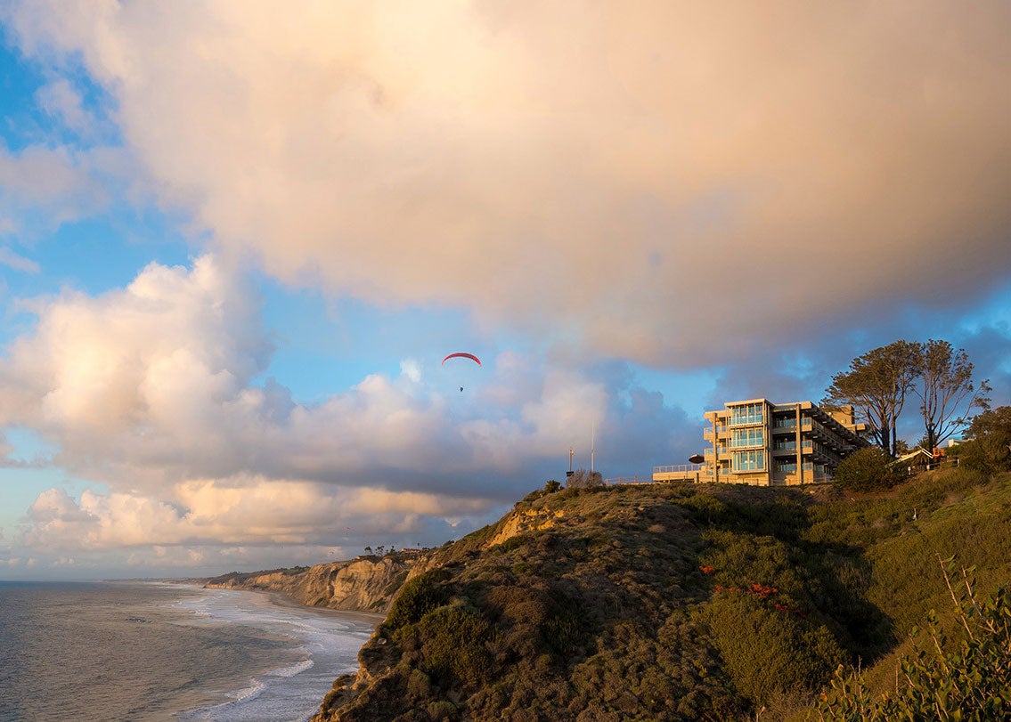 A coastal university building perched on a bluff above the ocean at sunset, with waves below, dramatic clouds overhead, and a paraglider soaring in the sky.