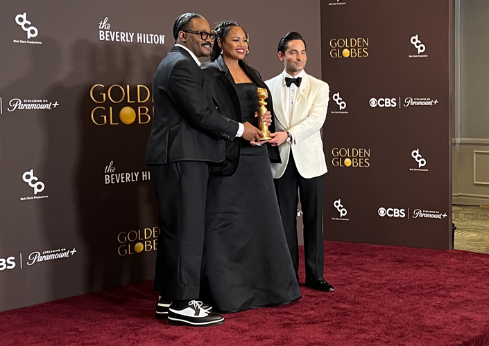 Three people in formal wear hold an award on a red carpet